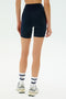 A person stands facing away, wearing SPLITS59 Ella High Waist Airweight Shorts in Indigo/Pirate Red, white socks with navy stripes, and white sneakers against a plain white background.