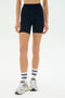 A person wearing SPLITS59 Ella High Waist Airweight Short in Indigo/Pirate Red, paired with white athletic socks and white sneakers, stands against a plain white background.