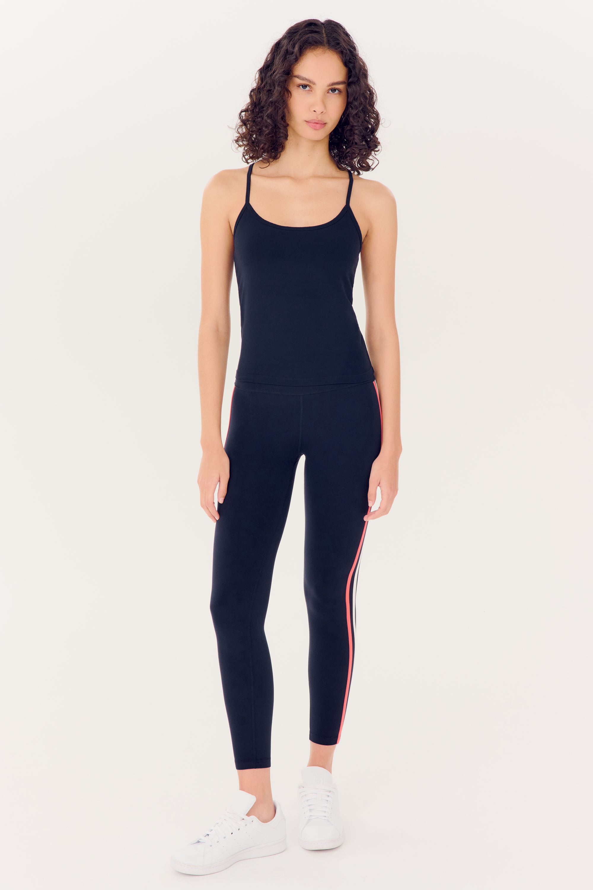 A woman in a black full-length SPLITS59 Airweight Tank and leggings with a red stripe stands against a white background, wearing white sneakers.