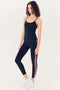 A young woman stands posing in a studio, wearing black activewear leggings and a full-length Airweight Tank in Indigo by SPLITS59 with stripe detail on the side, paired with white sneakers.