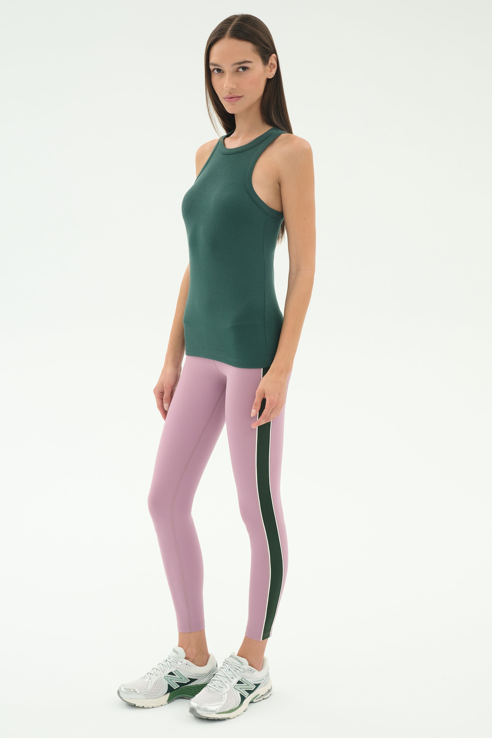 A woman stands against a plain background wearing the SPLITS59 Kiki Rib Tank Full Length in Military, light pink yoga leggings with a side stripe, and white athletic shoes—ideal for running or CrossFit workouts.