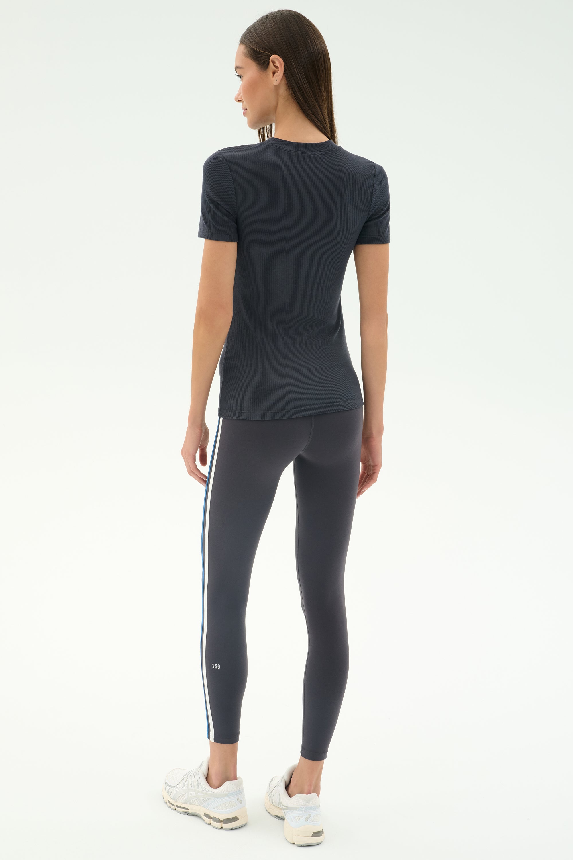 A woman faces away, wearing the SPLITS59 Louise Rib Short Sleeve in Graphite, dark leggings with side stripes, and white sneakers against a plain light background.