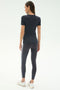 A woman faces away, wearing the SPLITS59 Louise Rib Short Sleeve in Graphite, dark leggings with side stripes, and white sneakers against a plain light background.