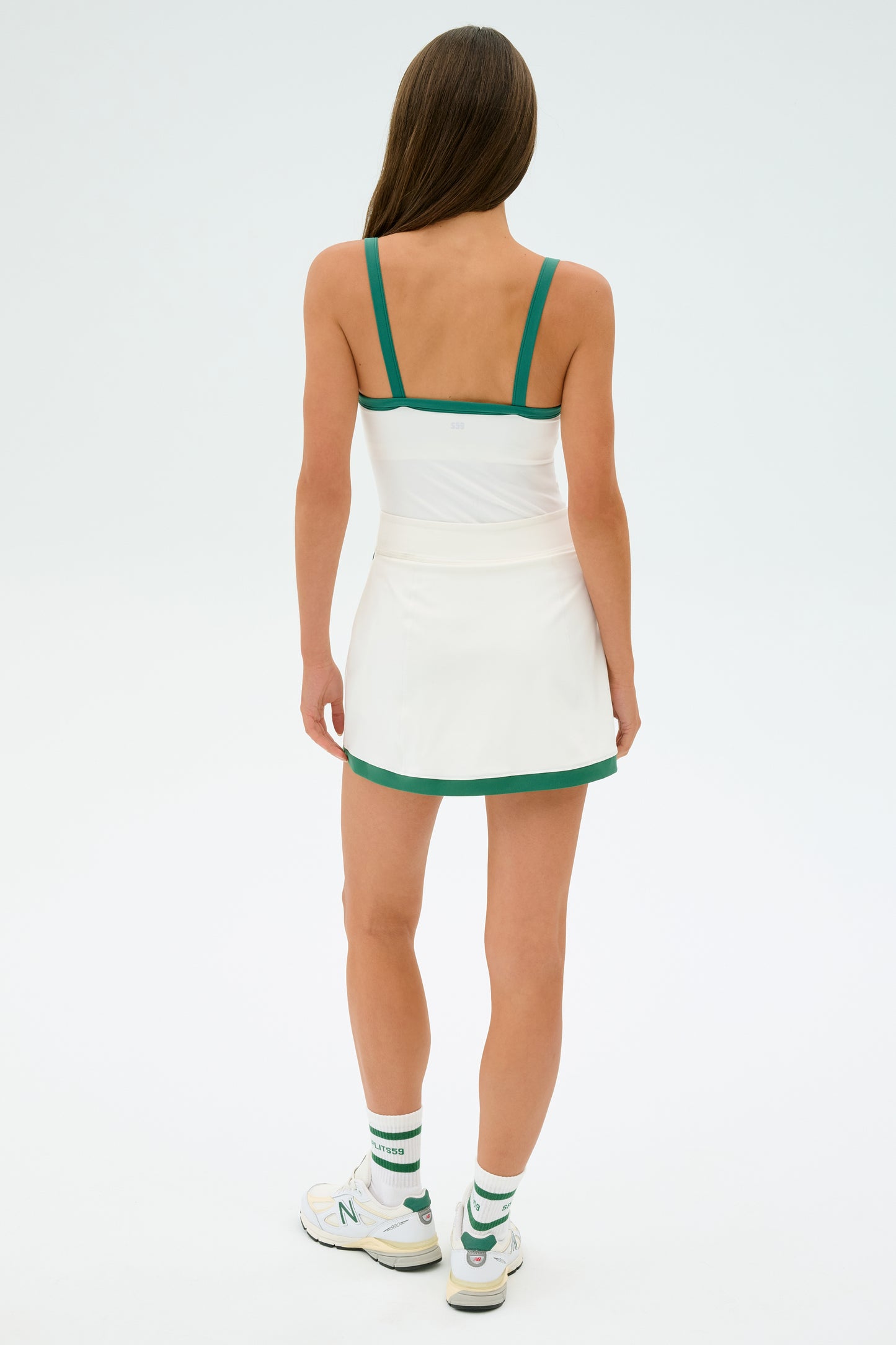 A person wearing the SPLITS59 Monah Rigor Tank in White/Arugula faces away, dressed in a white tennis outfit with green trim. They're also in white sneakers and socks with green lettering, set against a plain background.