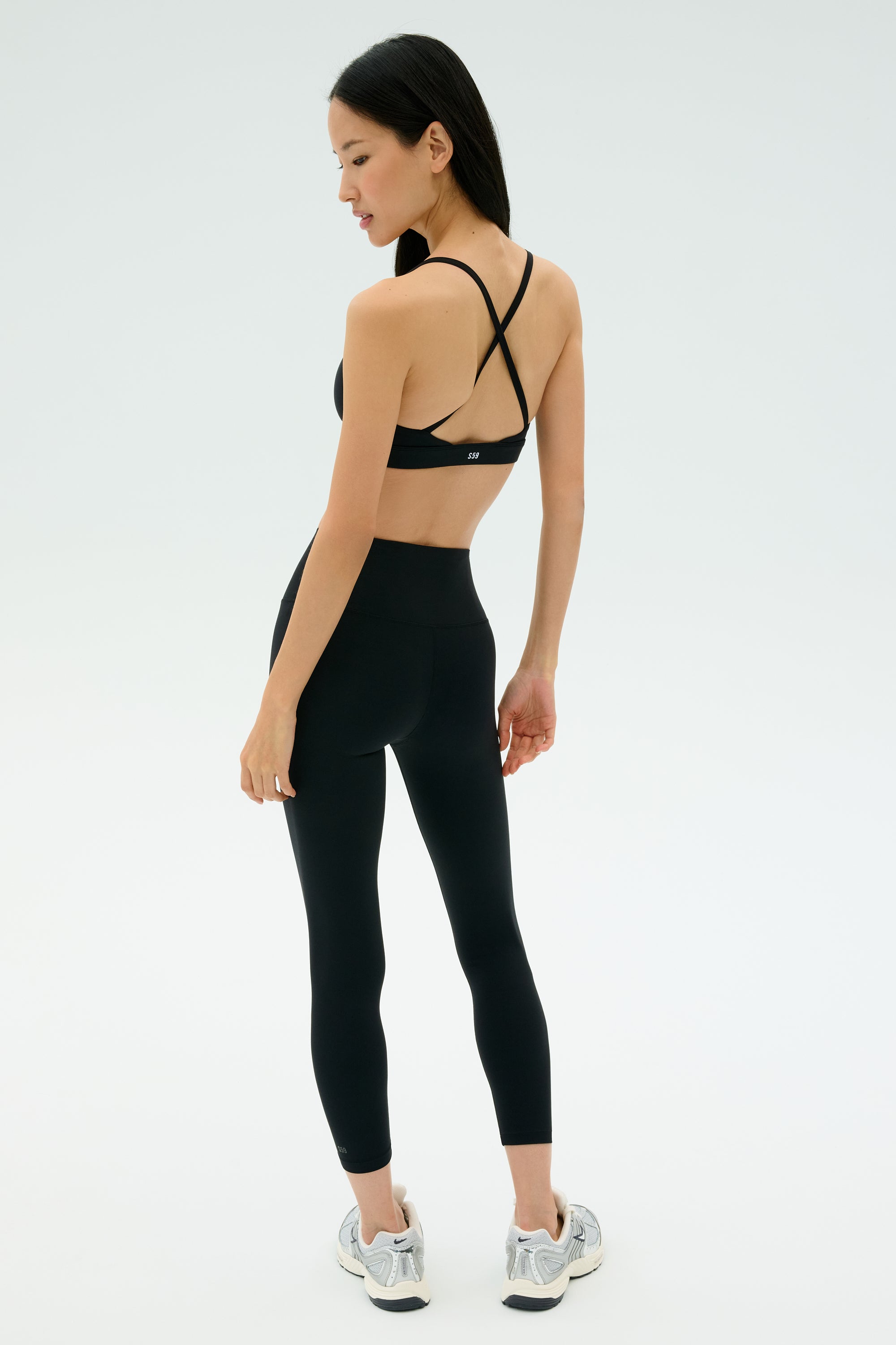 A woman wears the SPLITS59 Theo Rigor Bra - Black with black leggings and gray running shoes, standing and looking over her shoulder. Ideal for multi-sport performance, set against a plain background.