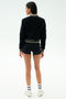 A person stands facing away in the SPLITS59 Willa Sherpa Jacket - Black, paired with black shorts, striped white socks, and white sneakers against a plain background—ideal for winter workouts.