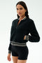 A woman stands against a plain background wearing the SPLITS59 Willa Sherpa Jacket in Black with striped cuffs and black fitted shorts—an ideal combo for winter workouts.