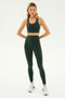 A person stands in a confident pose wearing a green sports bra, matching Airweight Super High Waist 7/8 leggings in the Military color by SPLITS59, and white sneakers against a plain background.
