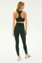 A woman in a dark green athletic outfit stands facing away, wearing a sports bra, white sneakers, and the SPLITS59 Airweight Super High Waist 7/8 leggings in Military. She's in a neutral, well-lit environment perfect for hot yoga.