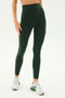 A person dressed in dark green Airweight Super High Waist 7/8 leggings from SPLITS59 and white-green sneakers stands against a plain background. The ultra luxe fabric of the leggings exudes a sense of elegance, while the person's left arm rests at their side.