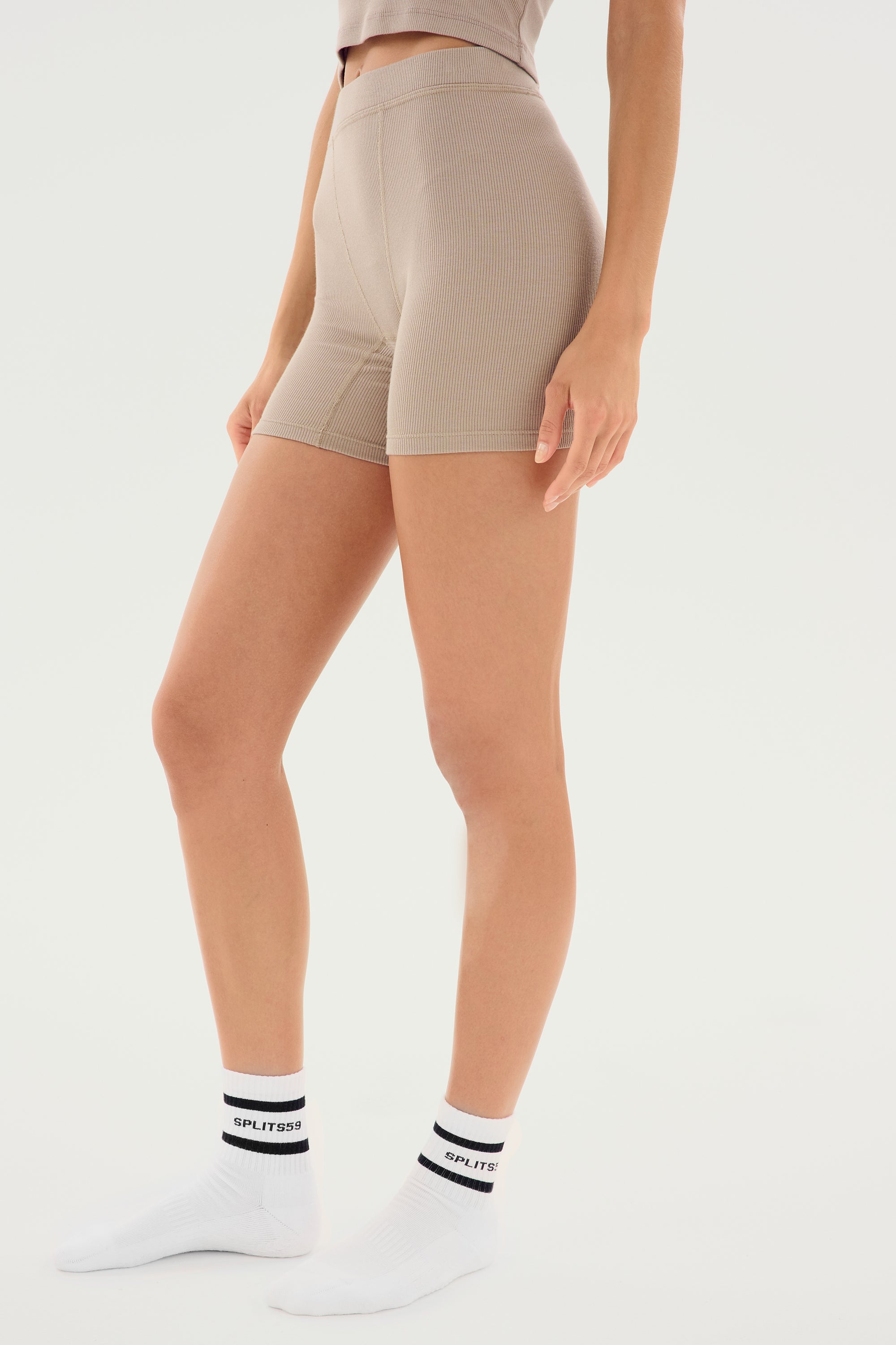 A person wearing Theo Rib Short - Latte from SPLITS59 in beige high-waisted style and white ankle socks with black writing stands against a white background, showcasing a classic design that exudes tailored comfort.