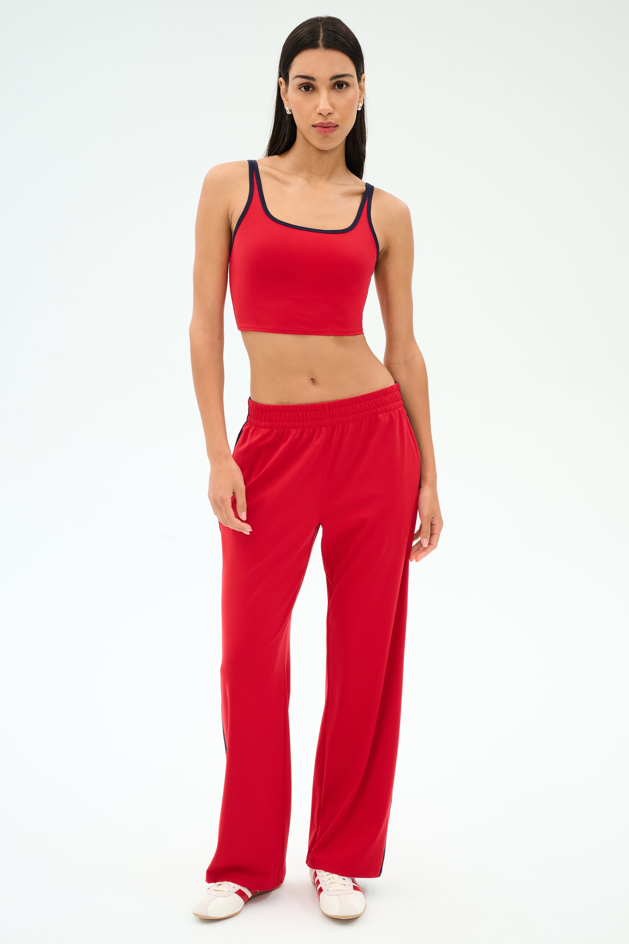 A woman stands against a plain background wearing a red crop top, SPLITS59 Maxie Rigor Track Pant in Pirate Red/Indigo, and white sneakers.