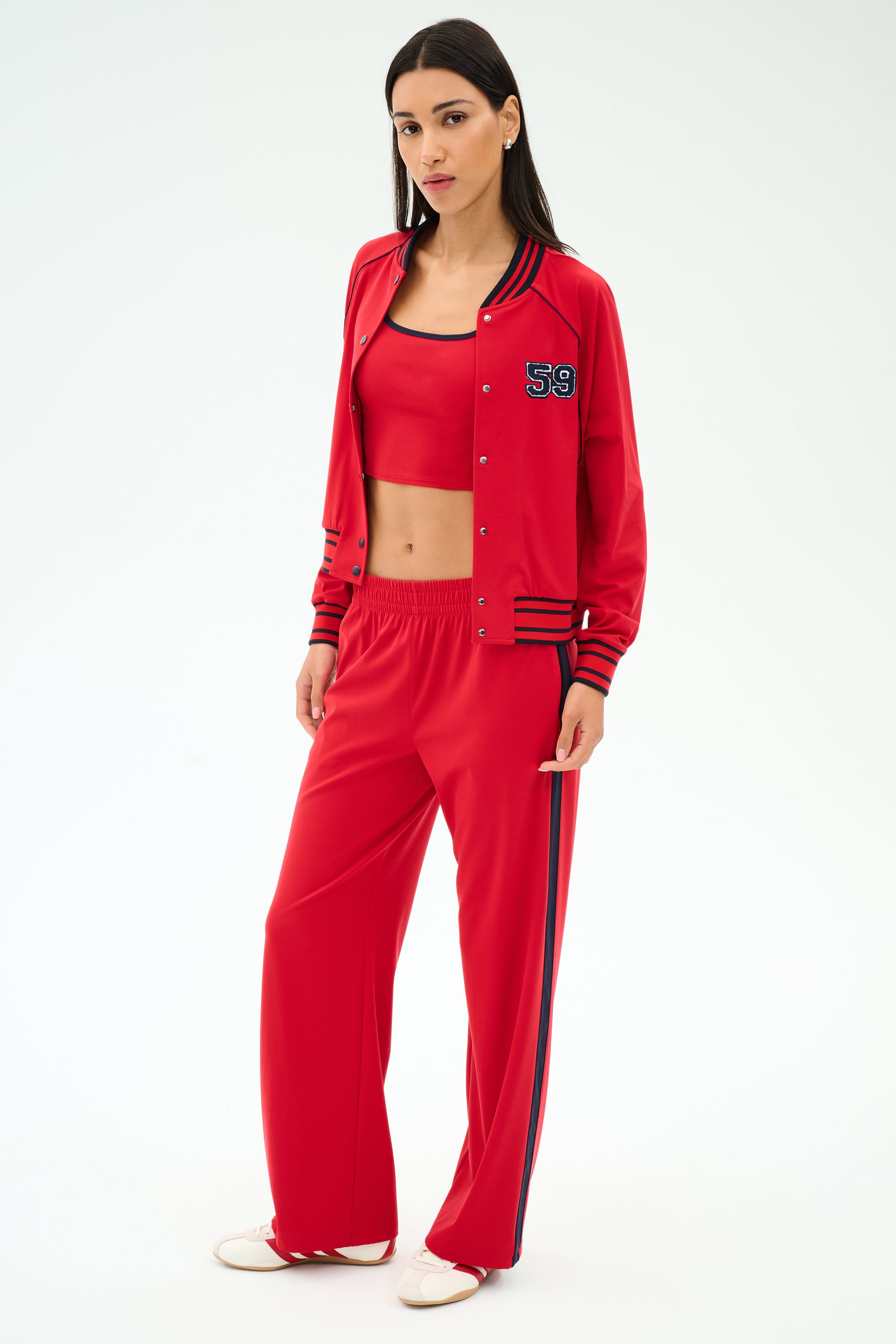 A person stands against a plain background wearing the SPLITS59 Hayes Varsity Rigor Jacket in Pirate Red, paired with a matching crop top and pants featuring black stripes, all crafted from stretch fabric.