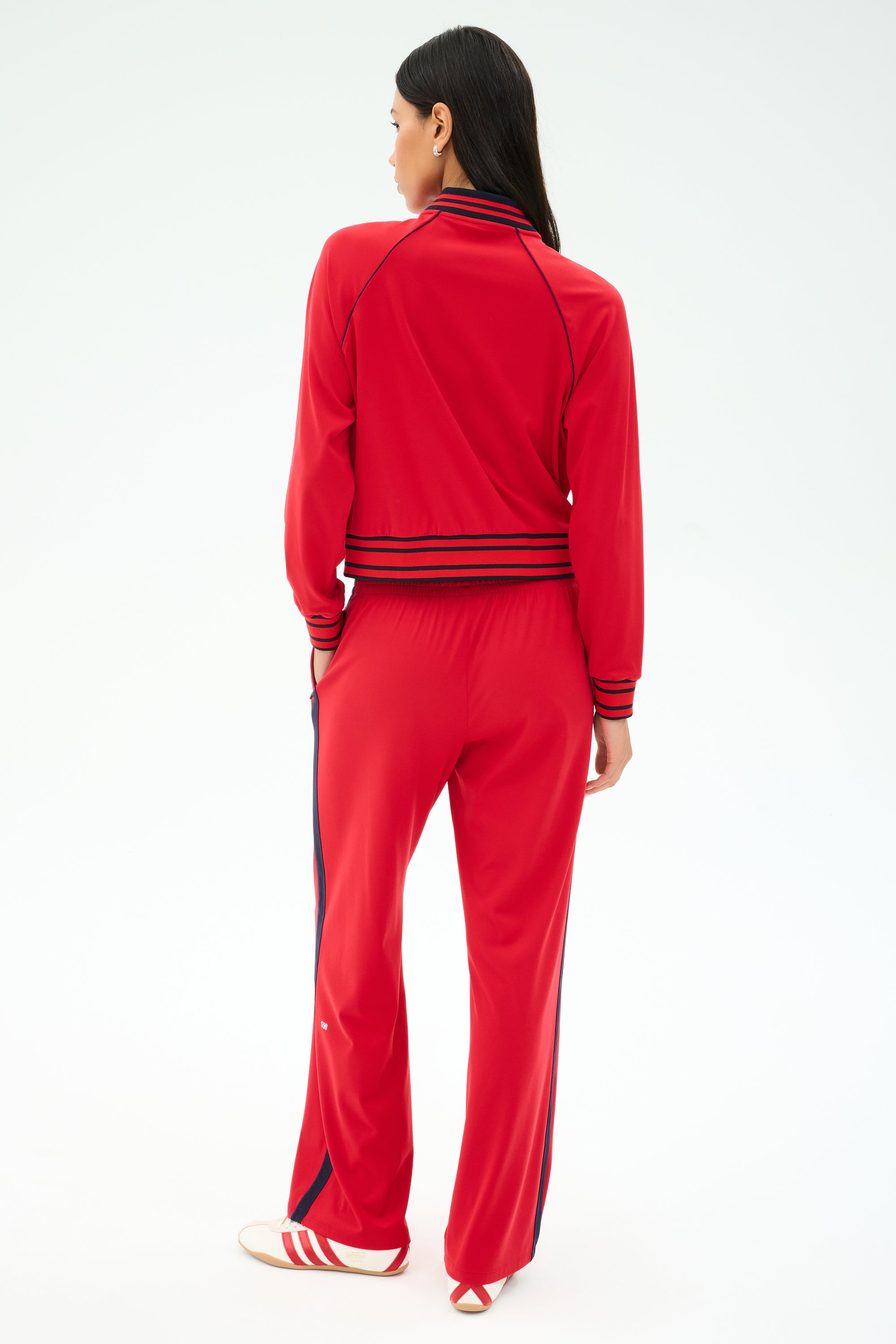 A woman faces away in the SPLITS59 Hayes Varsity Rigor Jacket in Pirate Red, styled with navy and white stripes and white sneakers with red accents, set against a light background.
