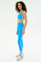 A woman wears the SPLITS59 Ella High Waist Airweight 7/8 leggings in bright blue with white stripes, paired with a matching sports bra, white socks, and gray sneakers against a plain white background.