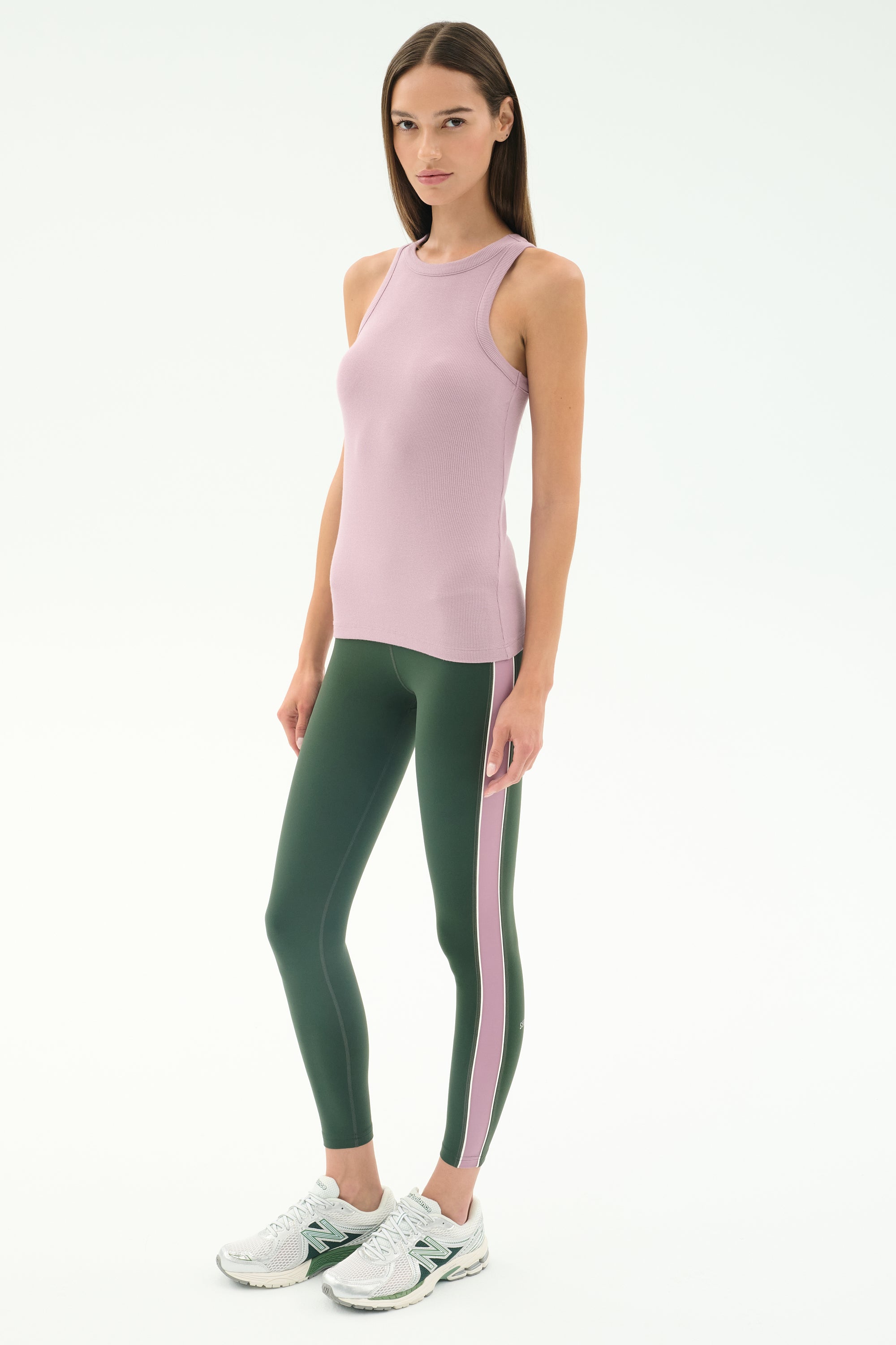 A woman poses against a plain background in the SPLITS59 Kiki Rib Tank Full Length - Blush, paired with green leggings featuring pink spandex stripes and white athletic shoes.