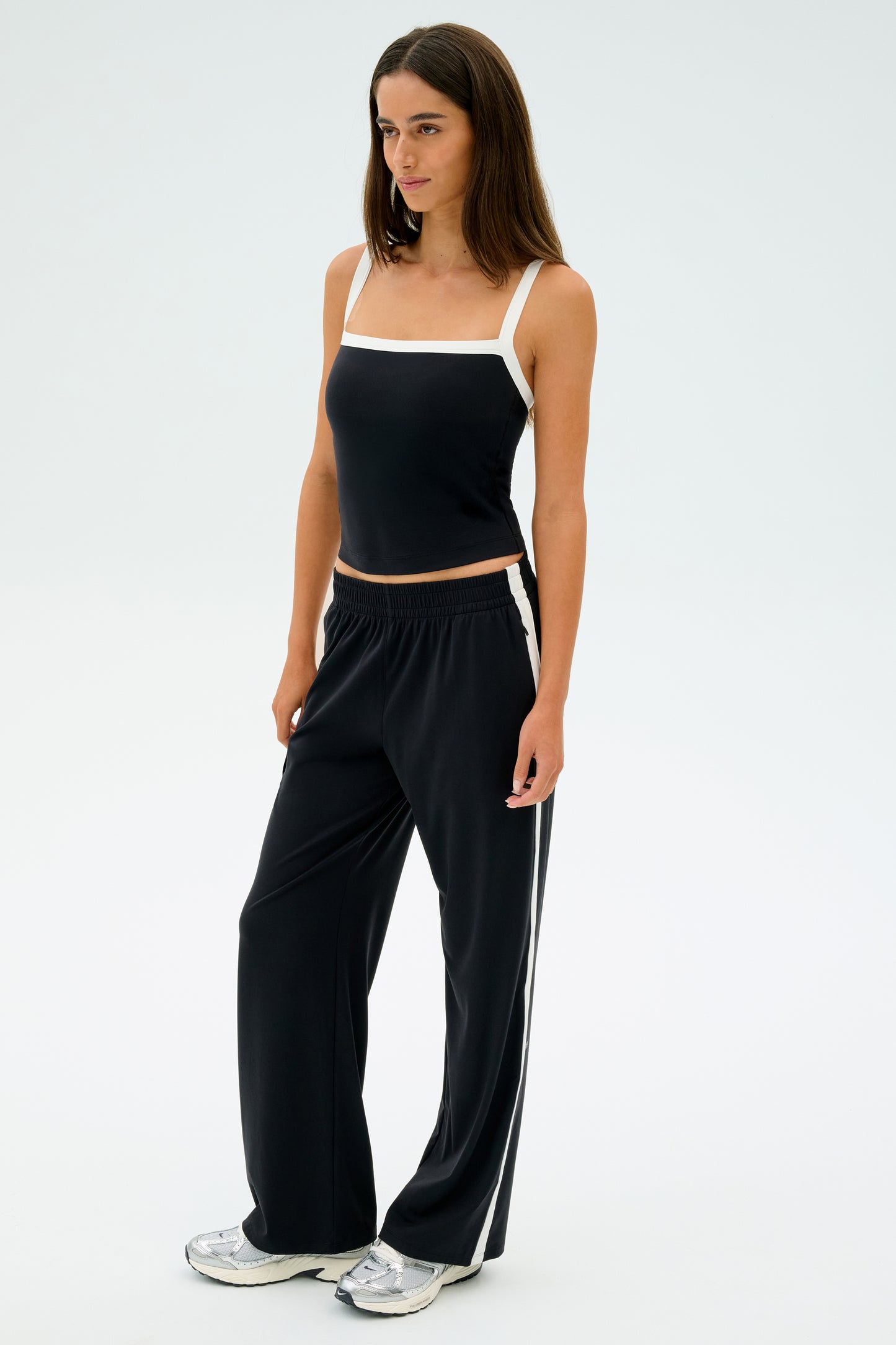 A woman poses against a plain white backdrop wearing the SPLITS59 Monah Rigor Tank in Black/White, black pants with white stripes, and silver sneakers.