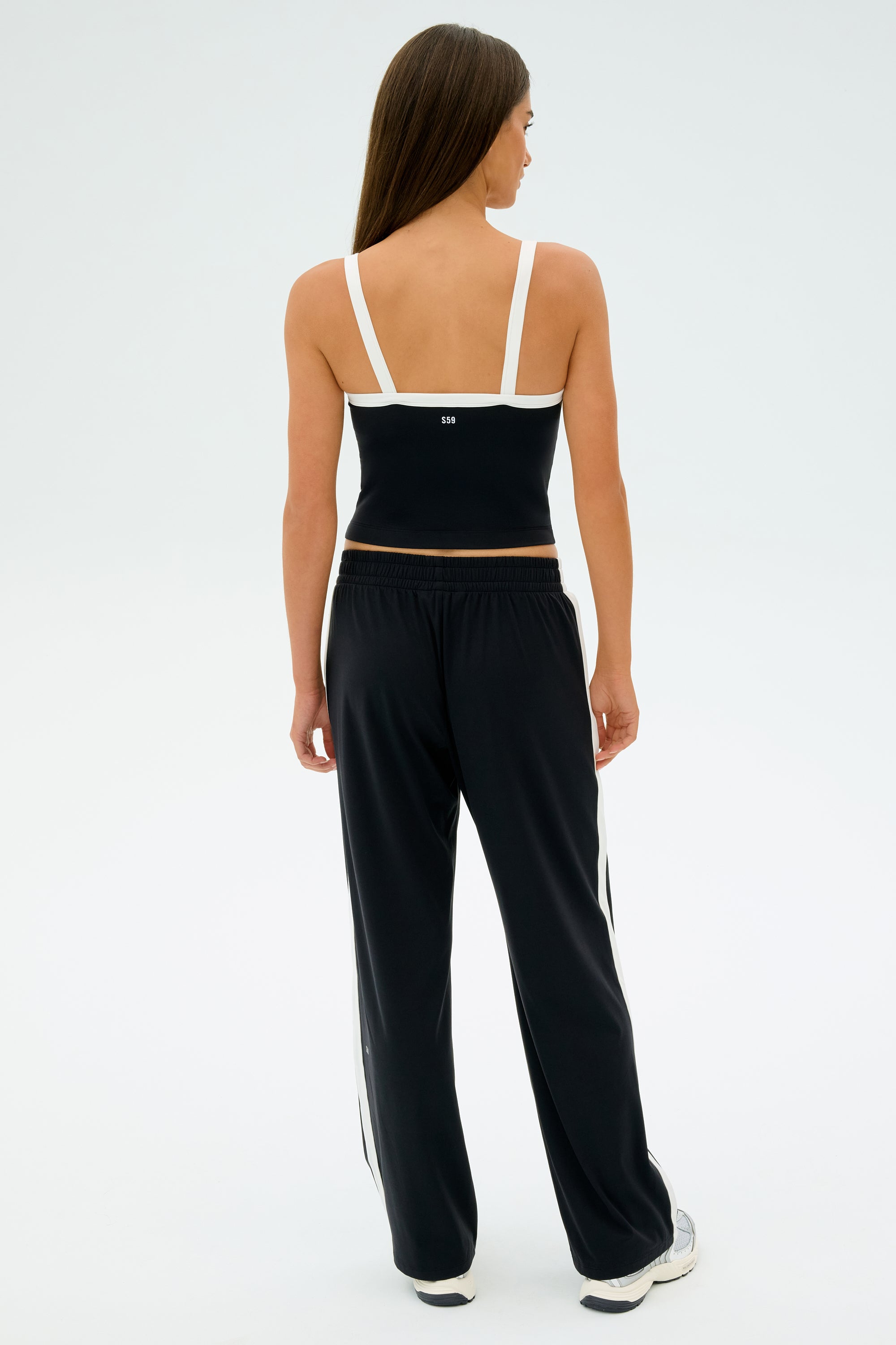 A woman with long hair is seen from behind wearing a SPLITS59 Monah Rigor Tank in black and white, paired with black pants, against a plain background.
