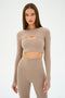 Wearing a beige long-sleeve crop top with a cut-out and matching high-waisted leggings, a person poses against a plain background, ready for yoga or dance. The SPLITS59 Amelia Airweight Lite Shrug in Latte adds extra style and comfort to their look.