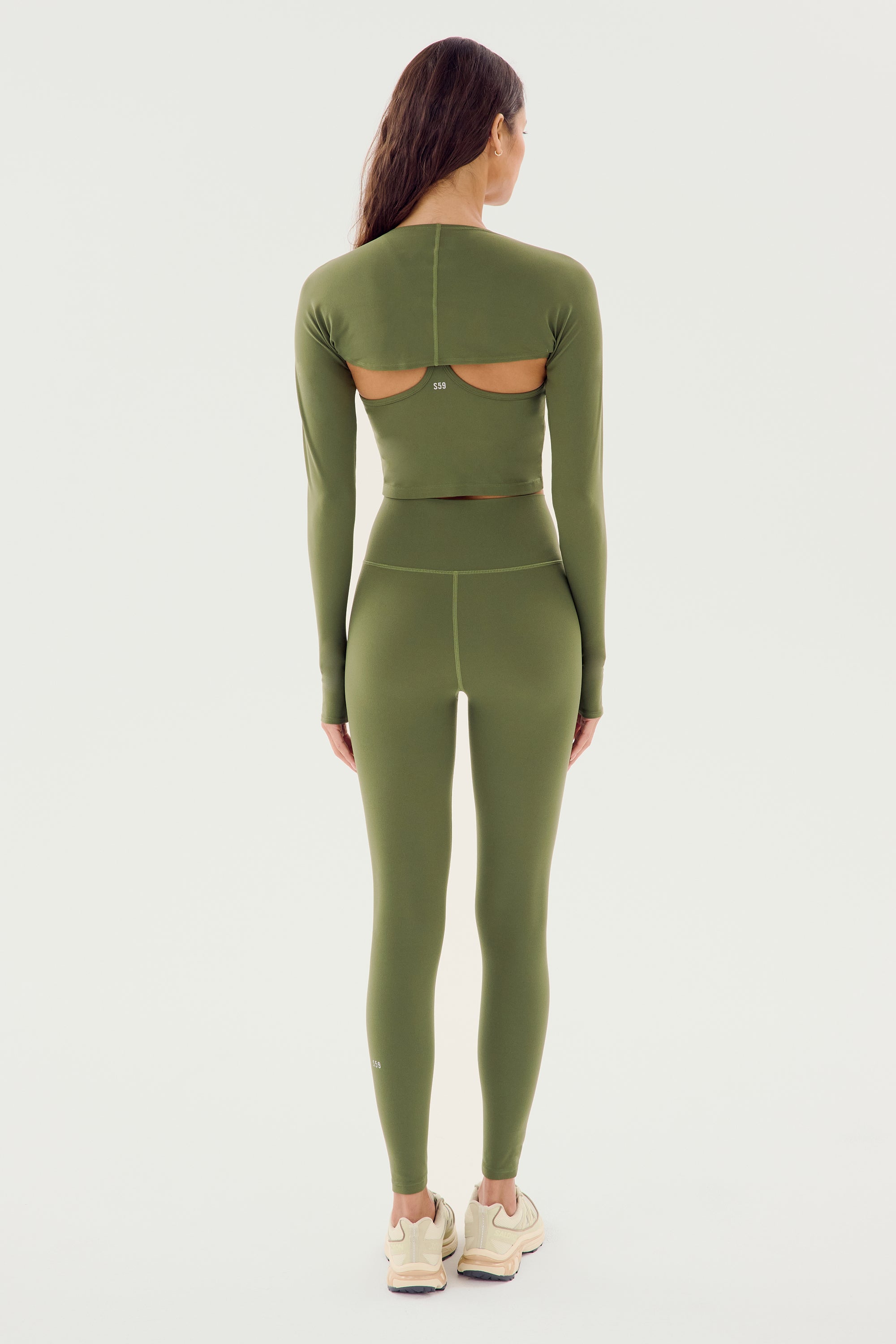 A person wearing SPLITS59's olive Airweight High Waist Legging and a long-sleeve, olive green athletic top crafted from a blend of nylon and spandex with cut-out back details, standing and facing away from the camera.