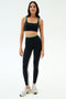 A woman wears the SPLITS59 Cait Rigor Bra in Black/Grass—black with green trim—paired with matching high-waisted leggings in Rigor fabric, both featuring a white side stripe, standing against a white background.