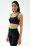 Woman wearing the SPLITS59 Cait Rigor Bra in Black/Grass, crafted from high performance Rigor fabric. Features supportive design ideal for high impact workouts. She stands against a plain light background, looking at the camera.