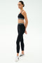 A woman stands in profile wearing the SPLITS59 Cali Active Rib Bra in black, paired with matching leggings featuring white stripes and white sneakers, set against a plain light background.