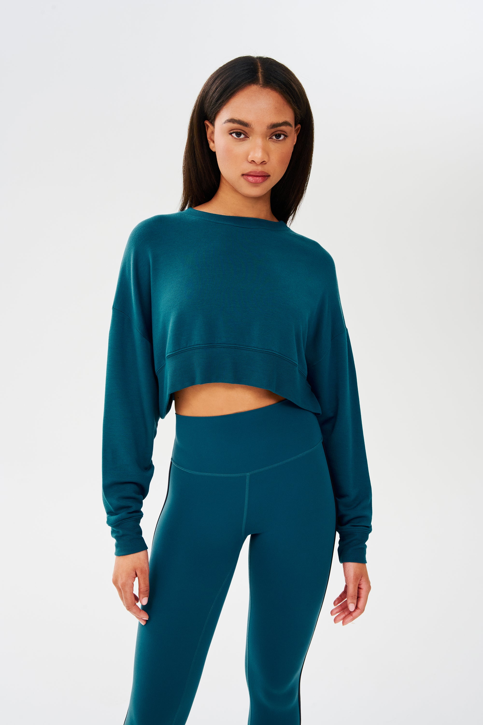 A woman wearing a SPLITS59 Noah Fleece Crop Sweatshirt in Peacock and leggings.