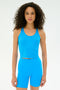 A woman with curly hair wears the SPLITS59 Ashby Rib Crop - Riviera, a bright blue sleeveless crop top, standing against a plain white background.