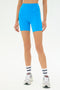 A person wearing SPLITS59 Ella High Waist Airweight Short in Riviera/White, paired with white socks and white athletic shoes, stands against a plain white background.