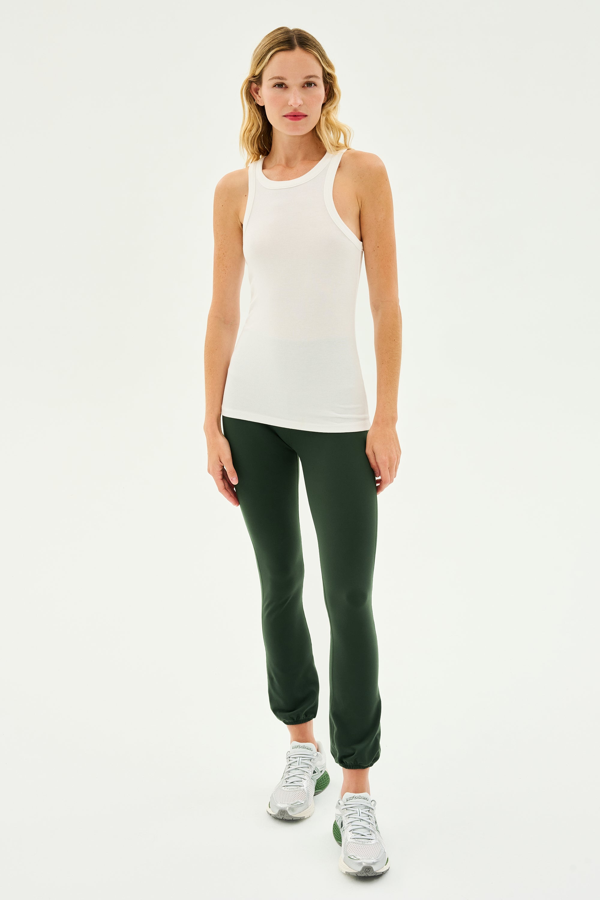 A woman stands facing forward wearing the Kiki Rib Tank Full Length in White by SPLITS59, dark green leggings, and gray athletic shoes against a plain white background, ready for yoga or gym workouts.