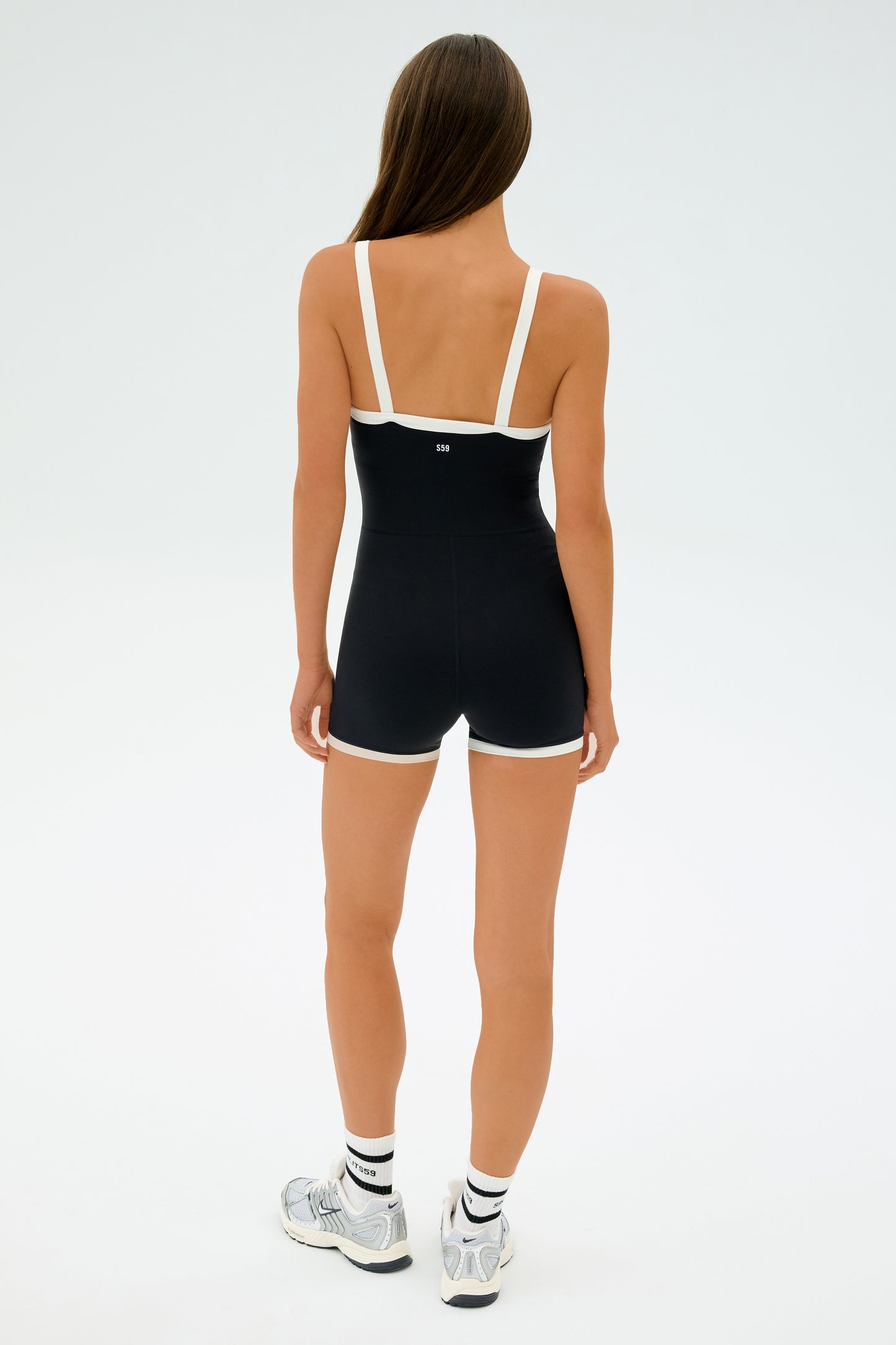 A person with long hair wears the SPLITS59 Monah Rigor Jumpsuit in black and white, paired with sleek white sneakers. They stand against a plain white background, facing away.