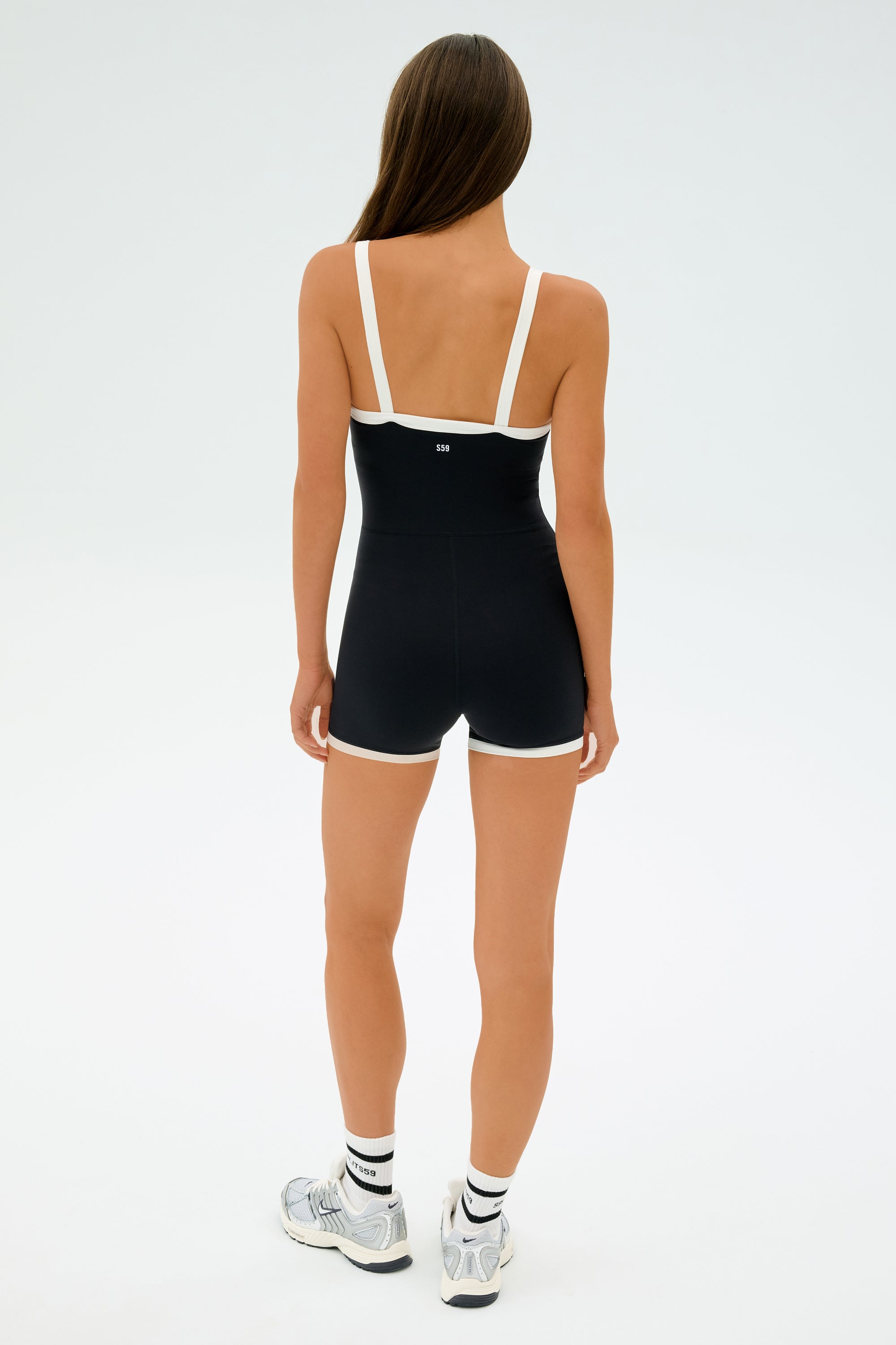 A person with long hair wears the SPLITS59 Monah Rigor Jumpsuit in black and white, paired with sleek white sneakers. They stand against a plain white background, facing away.