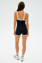 A person with long hair wears the SPLITS59 Monah Rigor Jumpsuit in black and white, paired with sleek white sneakers. They stand against a plain white background, facing away.