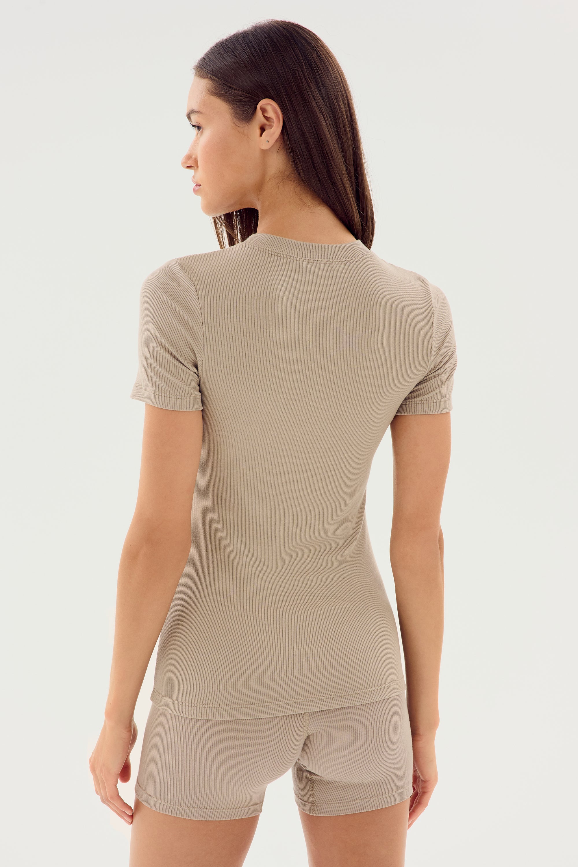 A person is standing and looking to the side, wearing the SPLITS59 Louise Rib Short Sleeve in Latte along with matching shorts made from a comfortable modal-spandex fabric. The photo is taken from behind.