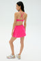 A woman faces away, wearing the SPLITS59 Venus High Waist Rigor Skort in Berry and a bright pink sports bra with silver and pink athletic shoes against a plain light background.
