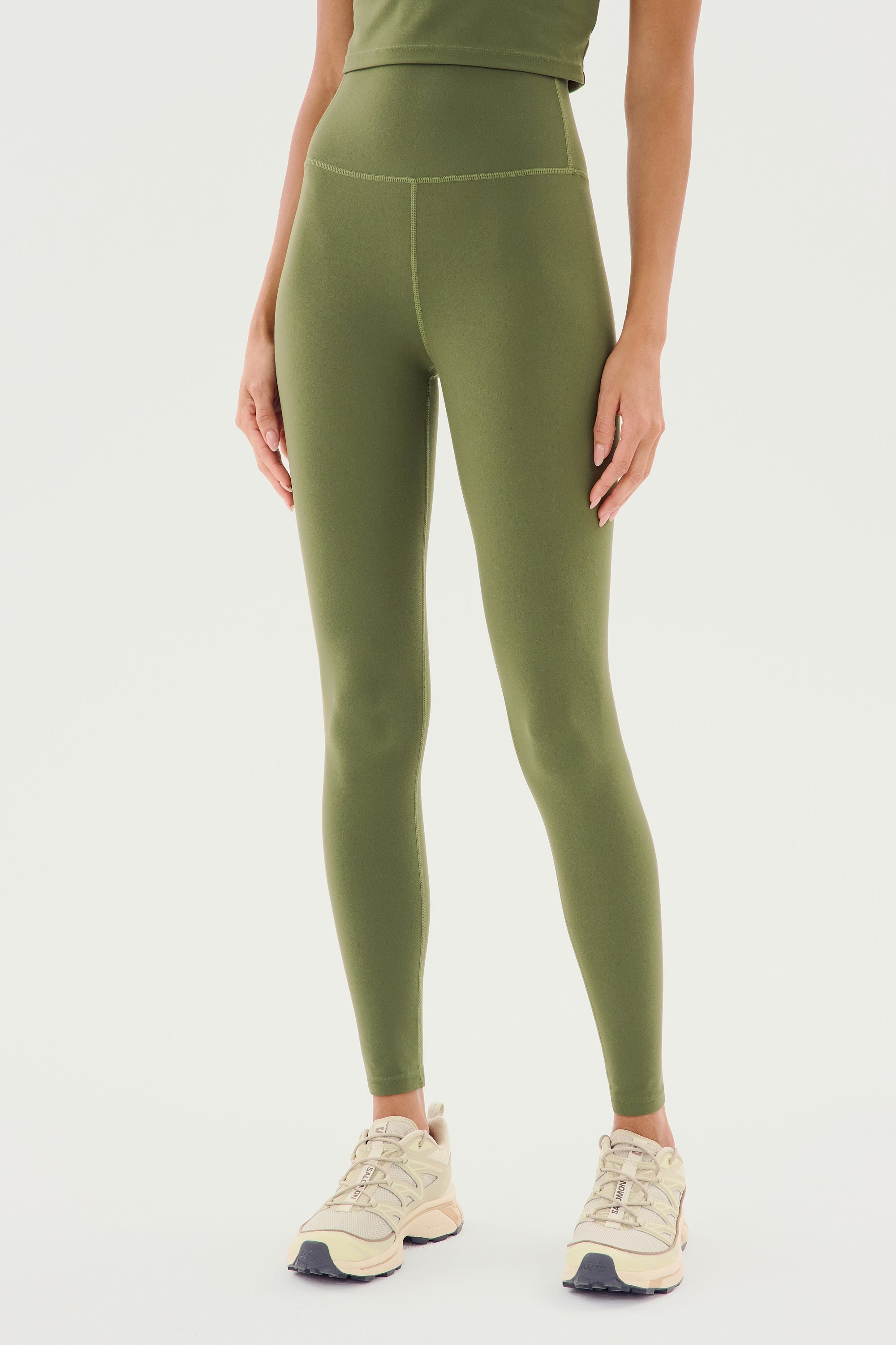 A person wearing SPLITS59's Airweight High Waist Legging in Olive, made from a comfortable nylon-spandex blend, paired with beige athletic shoes. The upper body is partially visible against a white background.