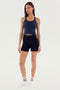 A woman stands wearing a navy blue SPLITS59 Ashby Rib Crop - Indigo, black shorts, and white socks against a plain white background.