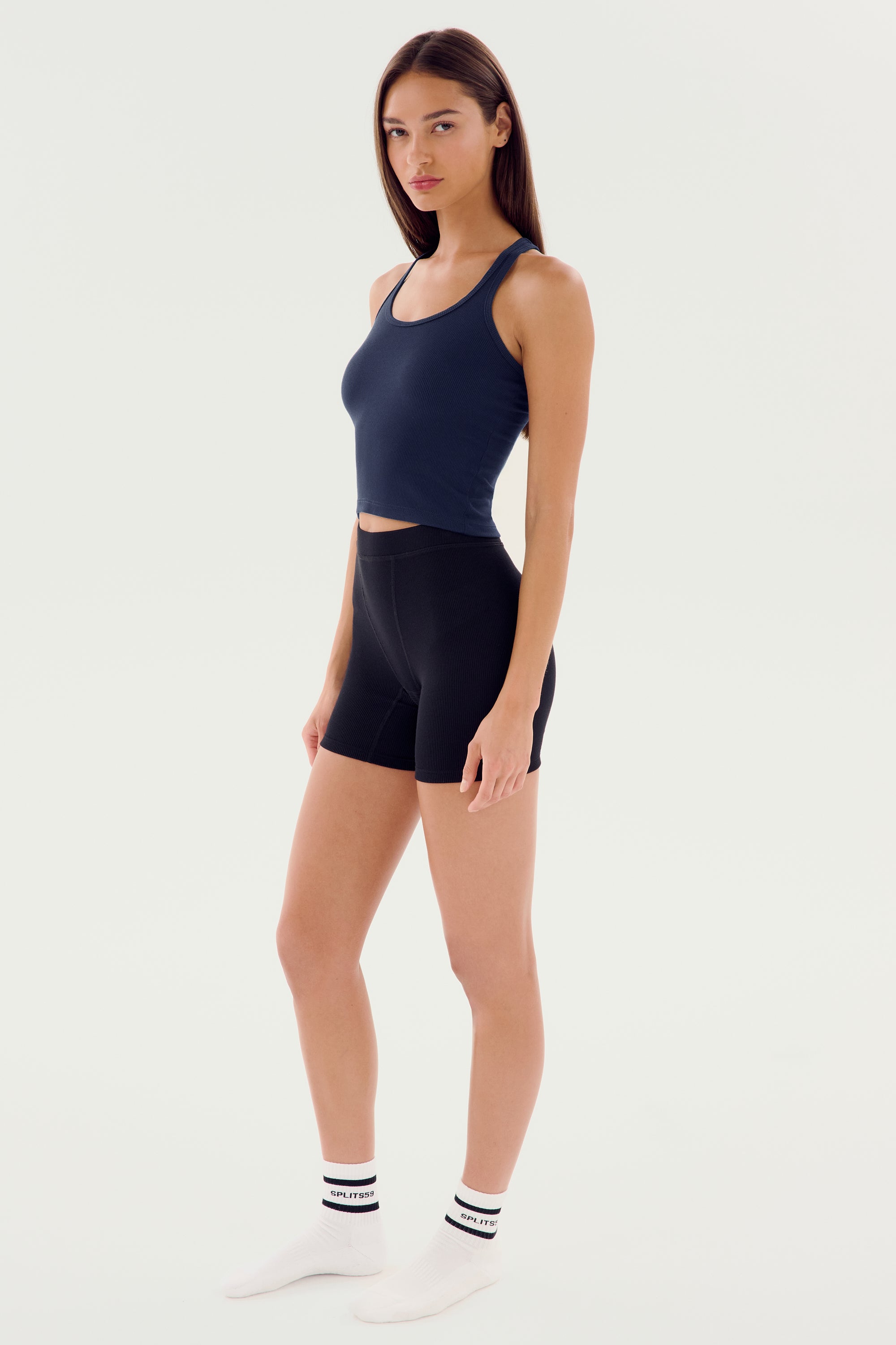 A person stands against a plain background, wearing a navy Ashby Rib Crop - Indigo by SPLITS59, black biker shorts, white socks, and white shoes. They are looking towards the camera with a neutral expression.