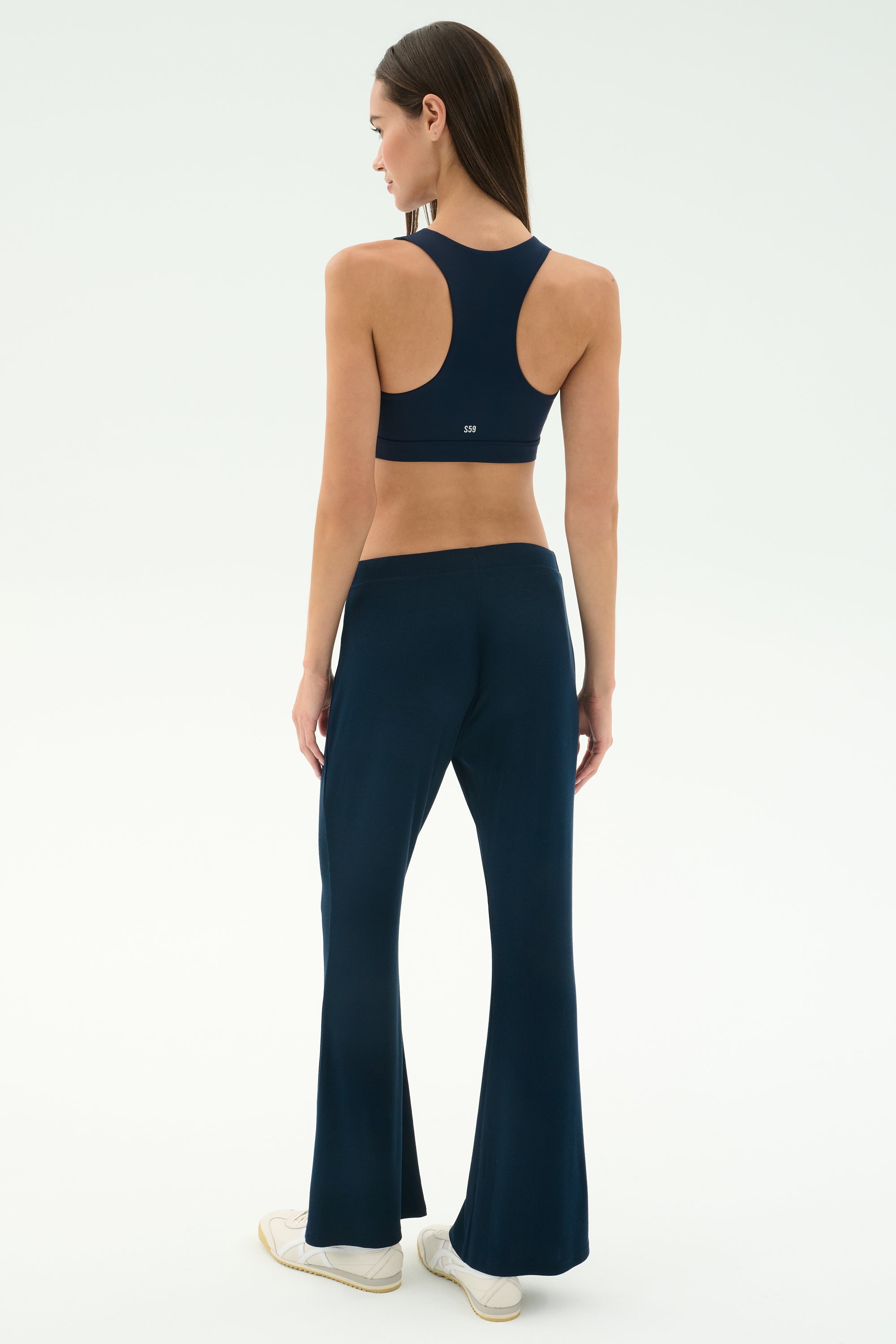 A woman faces away, wearing SPLITS59's Blaire Rib Pant in Indigo and a matching sports bra, both in soft modal fabric, with white sneakers against a plain light background.