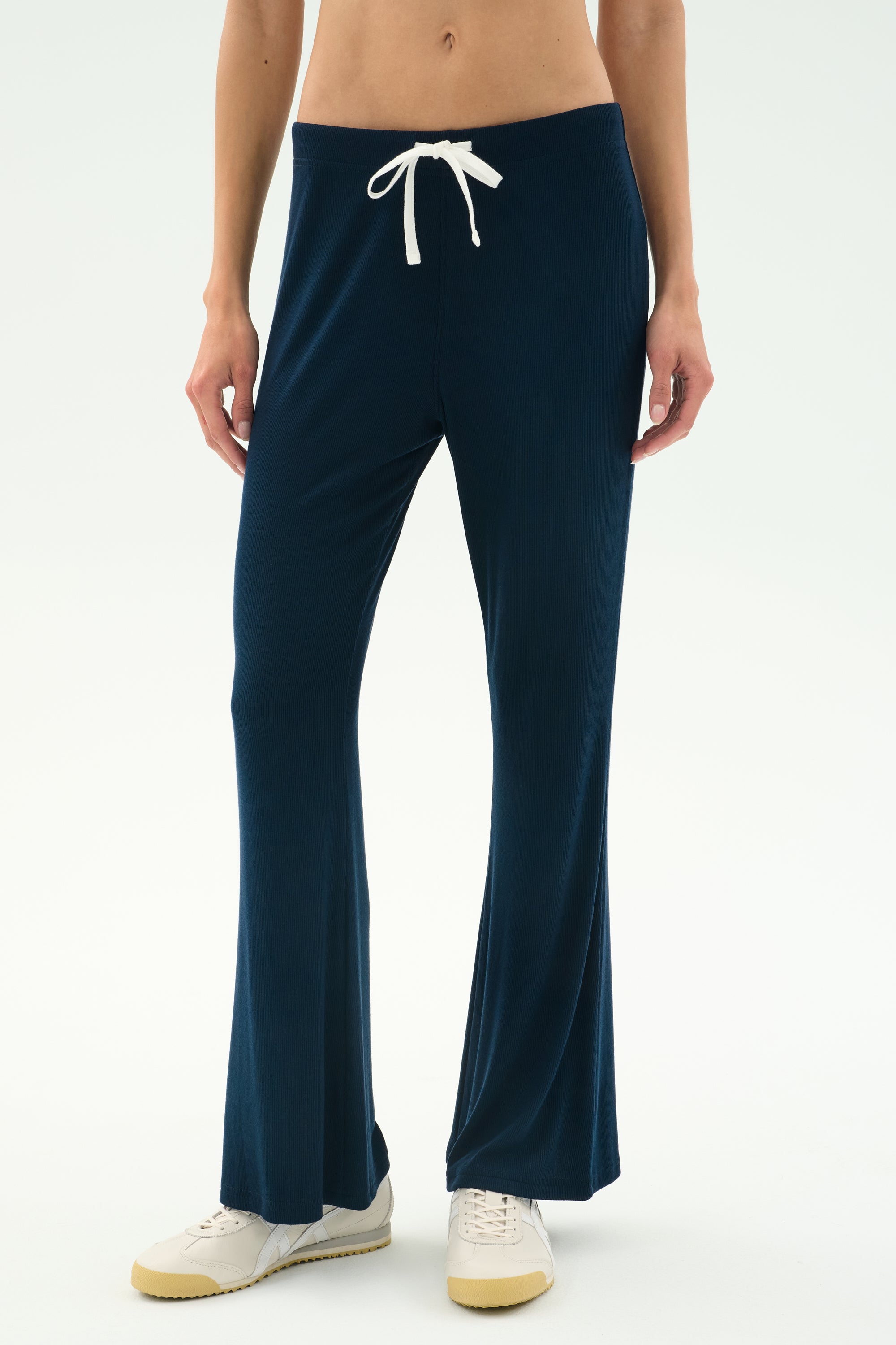 A person wears the SPLITS59 Blaire Rib Pant in Indigo—navy blue drawstring lounge pants crafted from soft modal with spandex—paired with white sneakers, standing against a plain light background.