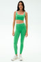 A woman faces forward, wearing the SPLITS59 Cait Rigor Bra in Grass/White with matching leggings and white sneakers, ready for high-impact workouts against a plain white background.