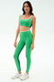 A woman stands against a plain background, wearing the SPLITS59 Cait Rigor Bra in Grass/White, paired with matching high-waisted leggings featuring a black stripe—ideal for high-impact workouts—and white sneakers.