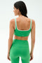 A woman faces away, wearing the SPLITS59 Cait Rigor Bra in Grass/White with matching high-waisted leggings, both crafted from Rigor fabric, against a plain light background.
