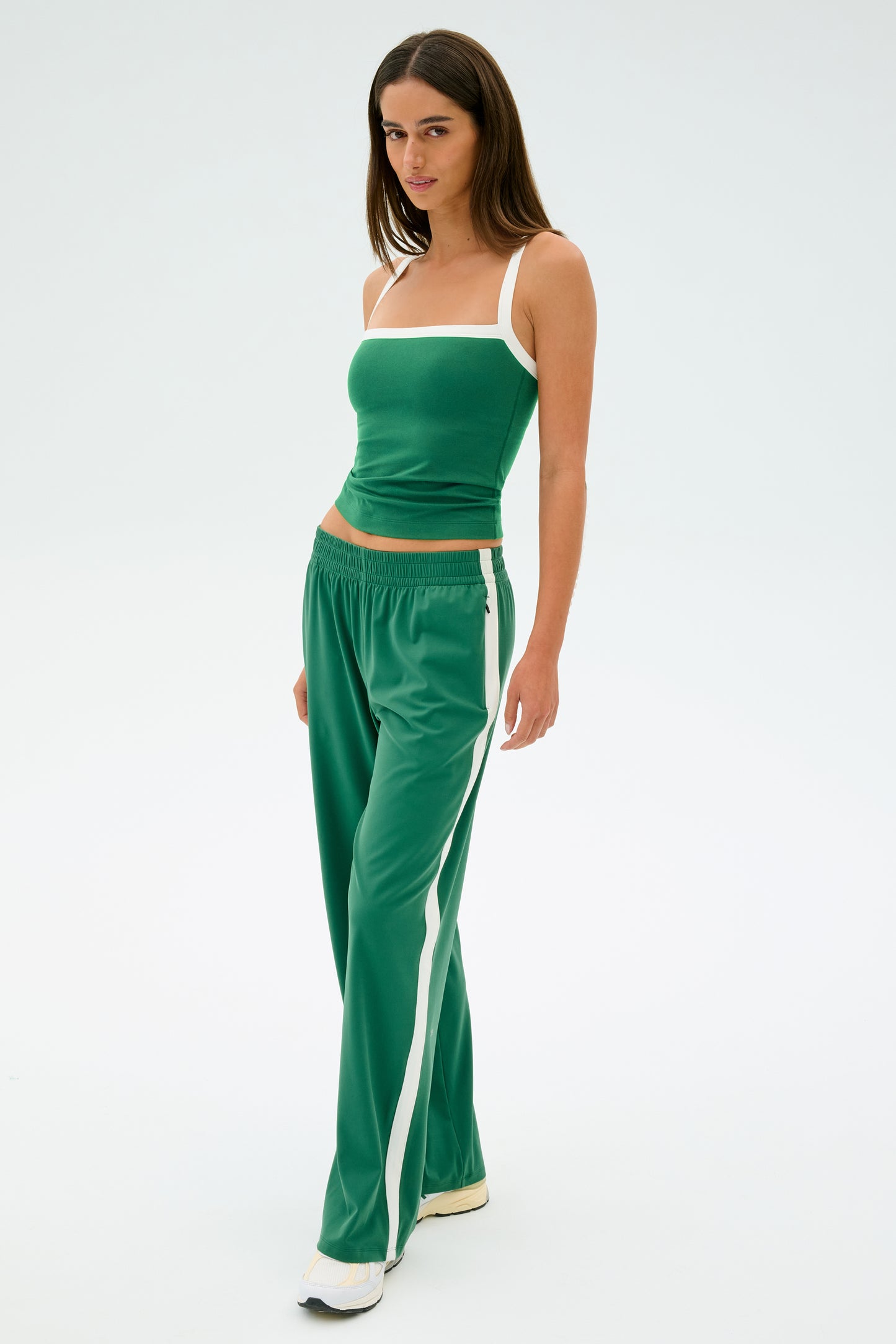 A person stands poised against a plain background, dressed in the Monah Rigor Tank in Arugula/White from SPLITS59, complemented by matching track pants featuring white stripes.
