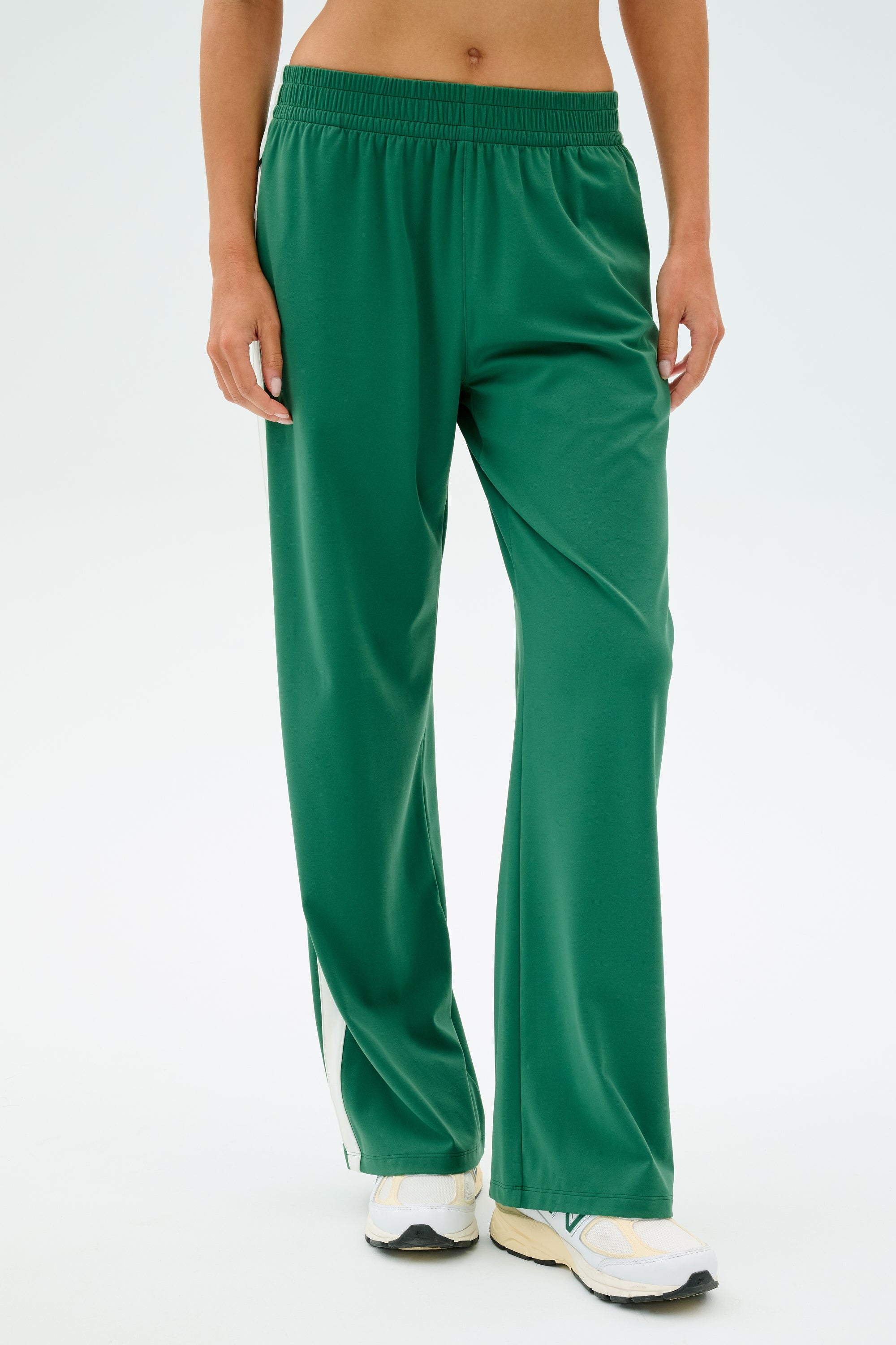 A person in SPLITS59's Maxie Rigor Track Pant in Arugula/White, featuring a white stripe and crafted from stretchable spandex, paired with white sneakers against a plain background.