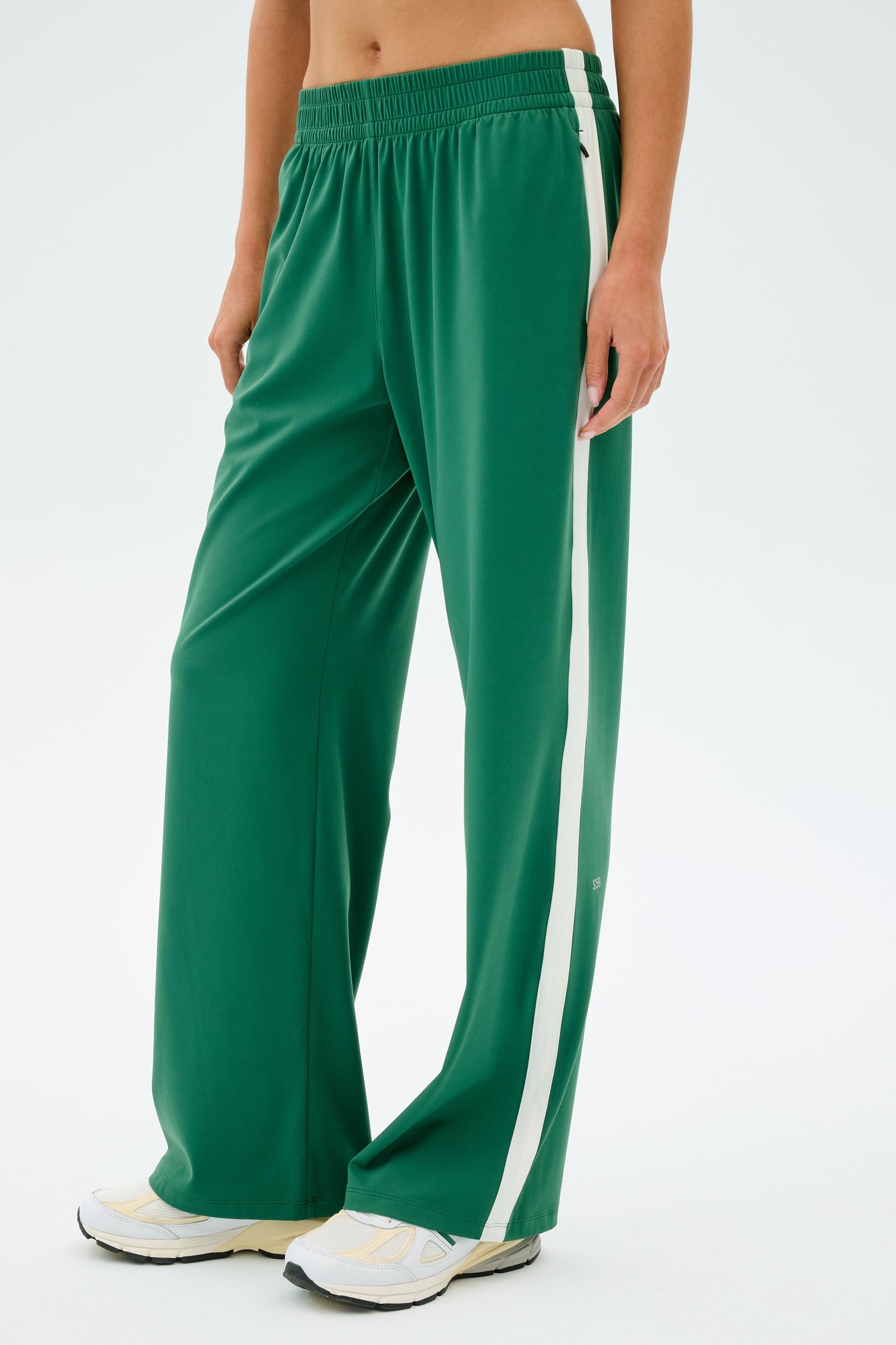 Someone in SPLITS59's Maxie Rigor Track Pant, featuring a white side stripe, paired with white sneakers stands against a plain background.