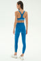 A woman faces away, wearing the SPLITS59 Ella High Waist Airweight 7/8 leggings in Stone Blue/Graphite with white and black side stripes, a matching sports bra, and white sneakers against a plain background—ideal for hot yoga.