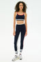 A woman faces forward against a white background, wearing the SPLITS59 Sam High Waist Rigor 7/8 leggings in Indigo/White with red and white stripes, paired with a matching sports bra, white socks, and athletic shoes.
