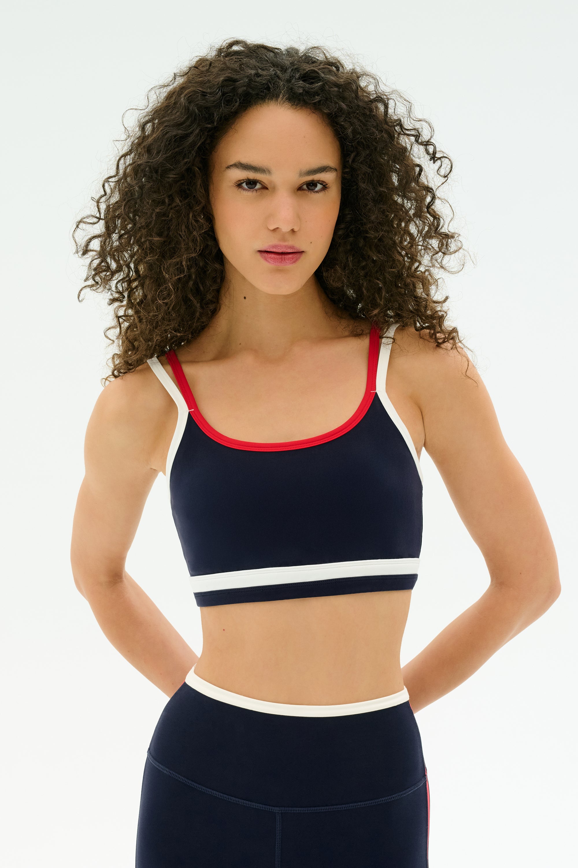 A woman with curly hair models the SPLITS59 Eva Rigor Double Strap Bra in Indigo/White and navy blue leggings with contrast details, standing against a plain white background, ready for gym workouts.